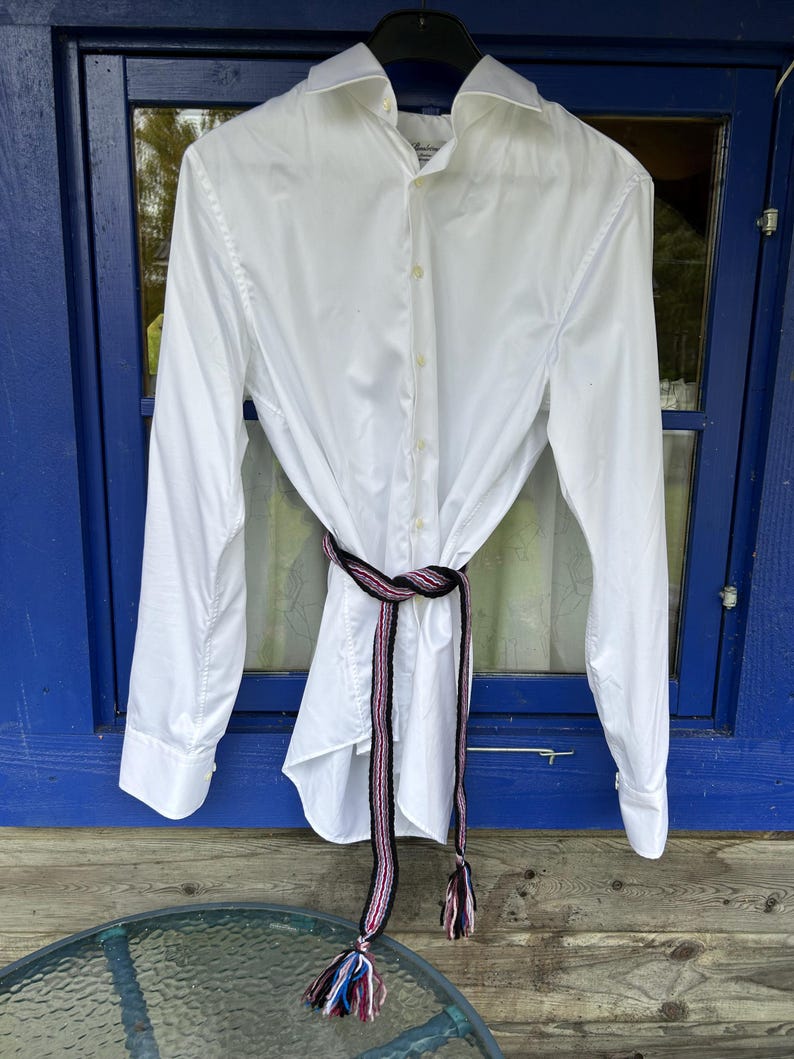 May include: A crisp white long-sleeved button-down shirt featuring a classic collar and cuffs. A long, narrow woven belt in black, pink, and white with multi-colored tasseled ends is tied around the waist of the shirt, creating a gathered effect.