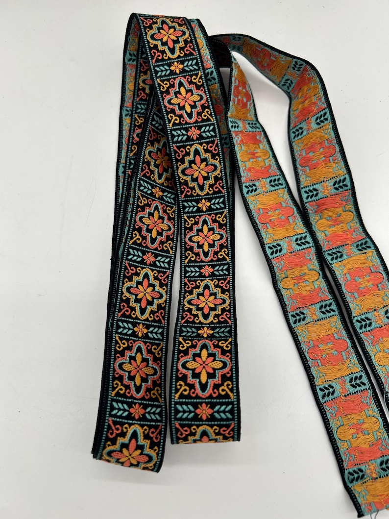 May include: Decorative ribbon with a black background and a repeating floral pattern in shades of orange, yellow, and teal. The ribbon is intricately designed with a detailed, symmetrical motif, suitable for various craft projects.