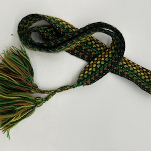 May include: A woven belt with a repeating pattern of green, yellow, and brown dots. The belt has a braided tassel at one end with green, yellow, and brown strands. The belt is on a white background.