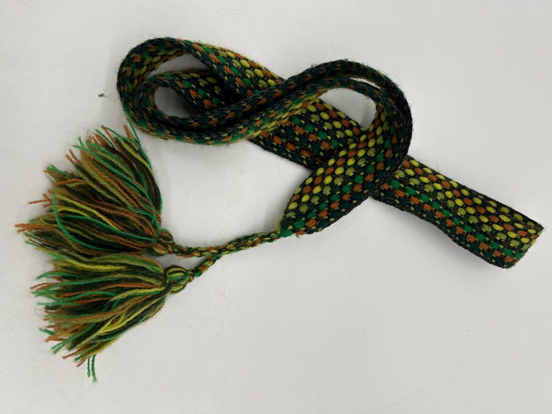 May include: A woven belt with a repeating pattern of green, yellow, and brown dots. The belt has a braided tassel at one end with green, yellow, and brown strands. The belt is on a white background.
