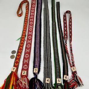 May include: Seven long, narrow woven fabric bands with intricate geometric patterns and fringe ends, numbered one through seven. The bands feature various color combinations including red, yellow, green, black, white, and grey, with two coins for scale.