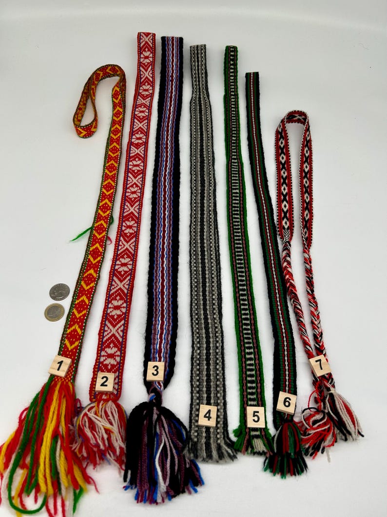 May include: Seven long, narrow woven fabric bands with intricate geometric patterns and fringe ends, numbered one through seven. The bands feature various color combinations including red, yellow, green, black, white, and grey, with two coins for scale.