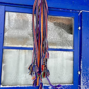 Pu&ograve; includere: Un tessuto colorato intrecciato con pi&ugrave; fili e nappe &egrave; appeso a un telaio di finestra blu. Il tessuto presenta una vivace miscela di strisce rosse, arancioni, viola e blu, con nappe decorative nella parte inferiore.