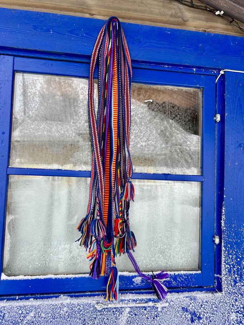 Pu&ograve; includere: Un tessuto colorato intrecciato con pi&ugrave; fili e nappe &egrave; appeso a un telaio di finestra blu. Il tessuto presenta una vivace miscela di strisce rosse, arancioni, viola e blu, con nappe decorative nella parte inferiore.