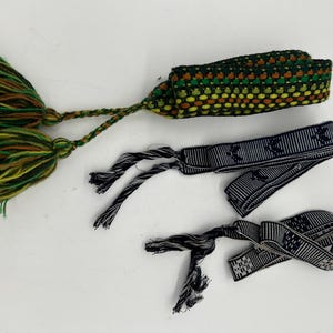 May include: Three handmade bracelets. One is a woven band in green, yellow, and brown with a tassel. Two are navy blue and white striped bands with a repeating pattern and tassels. The bracelets are on a white surface.
