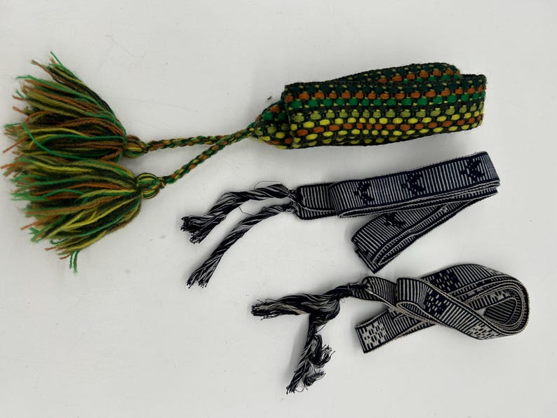 May include: Three handmade bracelets. One is a woven band in green, yellow, and brown with a tassel. Two are navy blue and white striped bands with a repeating pattern and tassels. The bracelets are on a white surface.