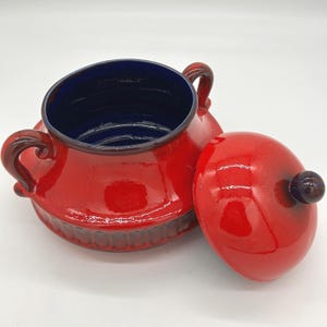 Puede incluir: Un azucarero de cerámica roja con tapa a juego. El cuenco tiene un interior azul oscuro y dos asas curvas. La tapa tiene un pequeño pomo morado oscuro. El exterior del cuenco es rojo brillante y el borde es de un tono más oscuro de rojo.