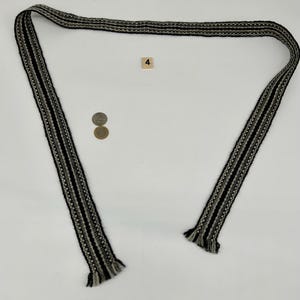 May include: A long, woven fabric belt or strap featuring a repeating pattern of black, white, and gray stripes. The ends of the strap are finished with short, frayed fringe. A small square tile with the number 4 and two coins are present.