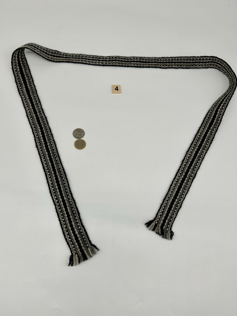 May include: A long, woven fabric belt or strap featuring a repeating pattern of black, white, and gray stripes. The ends of the strap are finished with short, frayed fringe. A small square tile with the number 4 and two coins are present.
