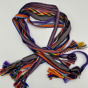 Pu&ograve; includere: Una cintura intrecciata multicolore con nappe. La cintura presenta diversi fili di vari colori, tra cui rosso, giallo, blu e viola. Le nappe sono attaccate alle estremit&agrave;. Probabilmente fatto a mano.