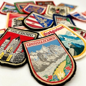 May include: A collection of embroidered souvenir patches featuring various Austrian landmarks and regional emblems. Notable patches include those labeled SALZBURG and GROSSGLOCKNER, depicting detailed mountain scenery, architectural structures, and colorful floral motifs with stitched borders.