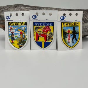 Op de afbeelding: Drie souvenirpatches met Zweedse ontwerpen. Eén patch toont een vrouw in traditionele klederdracht, een andere een rood Dala-paard en de derde een silhouet met de tekst "SWEDEN THE LAND OF MIDNIGHT SUN".