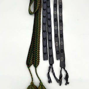 May include: Three woven belts with tassels. One belt is green, brown, and yellow with tassels. The other two are gray and black with geometric patterns. The belts are displayed against a white background.