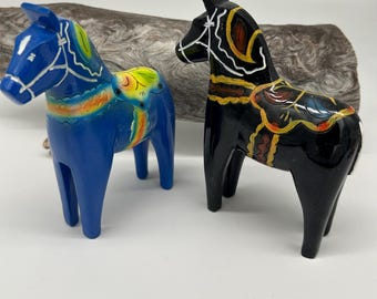 May include: Two wooden Dala horses. One is blue with yellow, orange, and green accents. The other is black with white, yellow, and red details. Both have painted saddles and bridles. They are standing on a white surface.