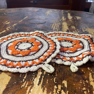 Puede incluir: Dos salvamanteles hechos a mano con ganchillo. Cada salvamanteles presenta un diseño floral naranja central rodeado de círculos concéntricos blancos, marrones y naranjas. Los bordes son festoneados. Estos salvamanteles decorativos son perfectos para la cocina.