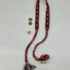 May include: A long, woven textile belt featuring a repeating diamond pattern in red, black, and white. The belt transitions into thick, braided cords at both ends, which terminate in loose, multi-colored fringe tassels. A small square tile with the number 7 is visible.