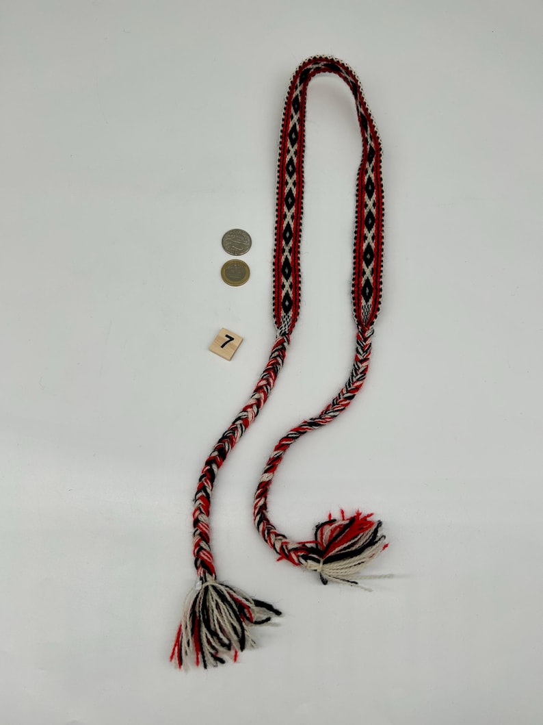 May include: A long, woven textile belt featuring a repeating diamond pattern in red, black, and white. The belt transitions into thick, braided cords at both ends, which terminate in loose, multi-colored fringe tassels. A small square tile with the number 7 is visible.