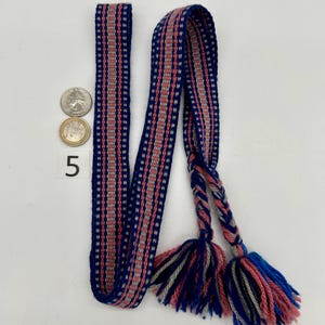 Pu&ograve; includere: Una cintura tessuta con sfondo blu e strisce rosa e grigie. La cintura ha nappe intrecciate in blu, rosa e grigio. Una moneta da venticinque centesimi e una moneta da un euro sono accanto alla cintura. Il numero 5 &egrave; stampato sulla superficie.