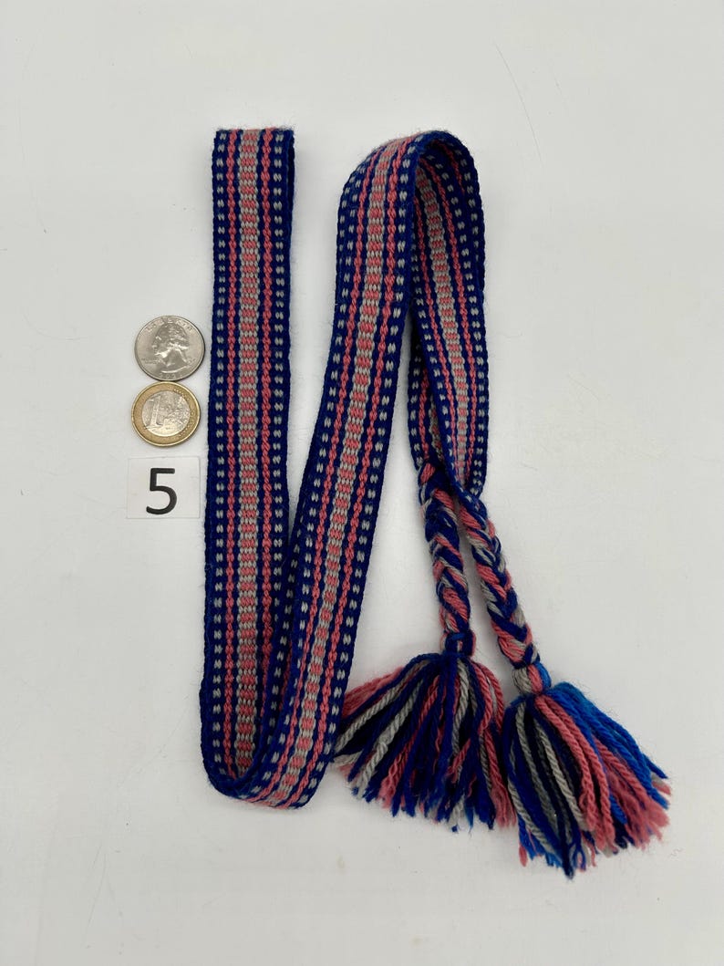 Pu&ograve; includere: Una cintura tessuta con sfondo blu e strisce rosa e grigie. La cintura ha nappe intrecciate in blu, rosa e grigio. Una moneta da venticinque centesimi e una moneta da un euro sono accanto alla cintura. Il numero 5 &egrave; stampato sulla superficie.