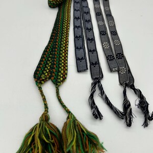 May include: Three woven textile straps with decorative tassels. One strap is primarily green, yellow, and brown with a patterned design. The other two are black and gray with repeating patterns. The tassels are green, yellow, and brown.