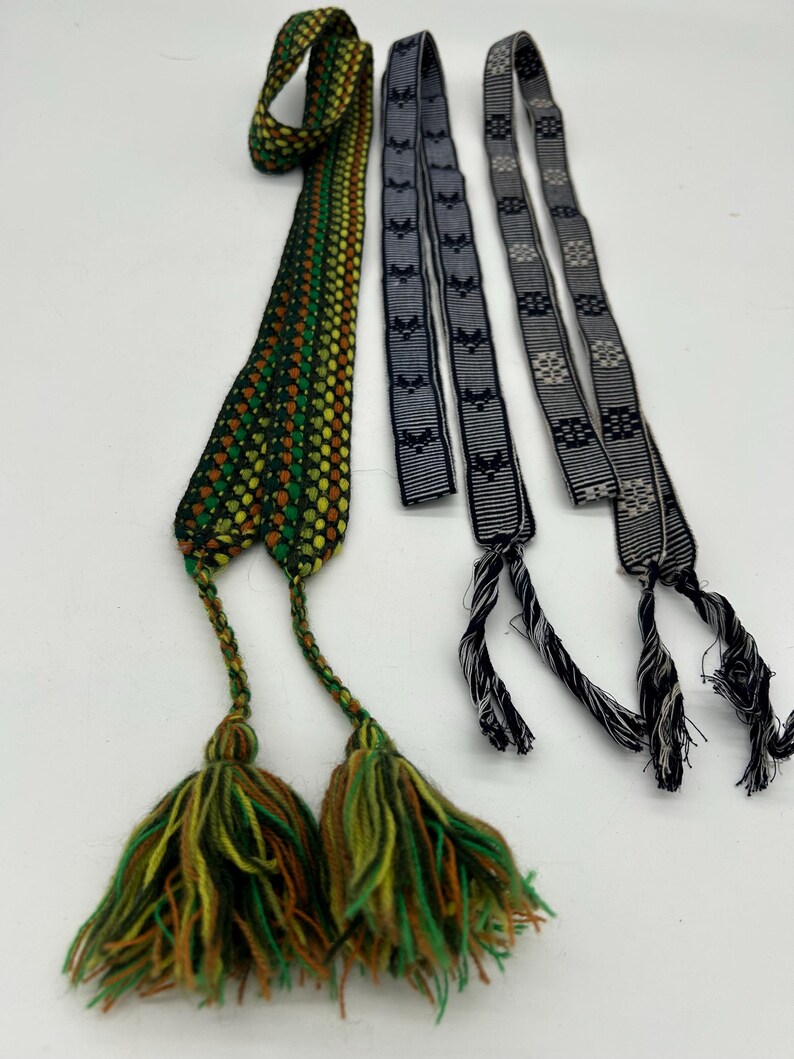 May include: Three woven textile straps with decorative tassels. One strap is primarily green, yellow, and brown with a patterned design. The other two are black and gray with repeating patterns. The tassels are green, yellow, and brown.