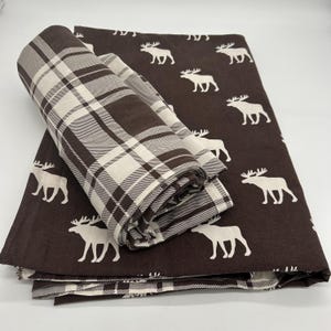 May include: A set of brown and white bedding. The flat sheet has a repeating pattern of white moose on a brown background. A rolled-up blanket and pillowcases with a brown and white plaid pattern are also visible.