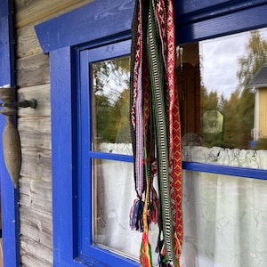 May include: A collection of long, narrow woven bands featuring intricate geometric patterns in vibrant colors including red, green, black, white, and yellow. Each band terminates in multi-colored yarn tassels hanging vertically from a blue window frame.