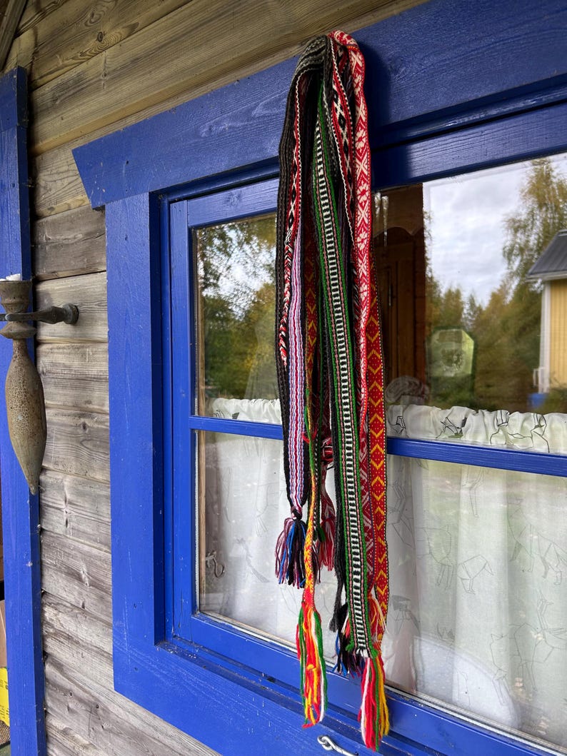 May include: A collection of long, narrow woven bands featuring intricate geometric patterns in vibrant colors including red, green, black, white, and yellow. Each band terminates in multi-colored yarn tassels hanging vertically from a blue window frame.
