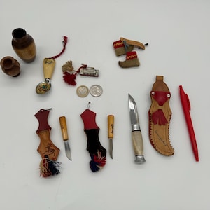 Può includere: Una collezione di piccoli oggetti fatti a mano, tra cui vasi di legno, coltelli decorativi con fodero in pelle, una penna e stivali in miniatura. I coltelli hanno manici in legno e lame in metallo. Alcuni oggetti hanno nappe.