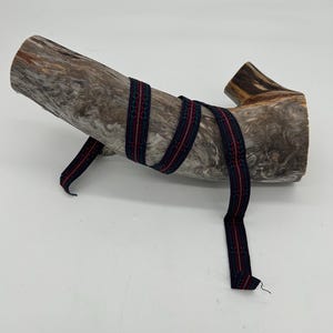 May include: A length of patterned ribbon in dark blue and red, wrapped around a piece of weathered wood. The ribbon features a repeating geometric design. The wood has a natural, textured appearance, with shades of brown and grey.