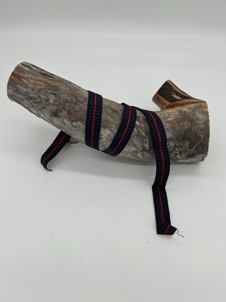 May include: A length of patterned ribbon in dark blue and red, wrapped around a piece of weathered wood. The ribbon features a repeating geometric design. The wood has a natural, textured appearance, with shades of brown and grey.