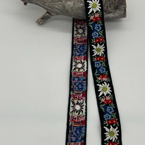 May include: Two decorative ribbons with floral patterns. One ribbon features a repeating pattern of colourful flowers on a black background. The other ribbon has a patchwork design with various colours and patterns. The ribbons are draped over a piece of driftwood.