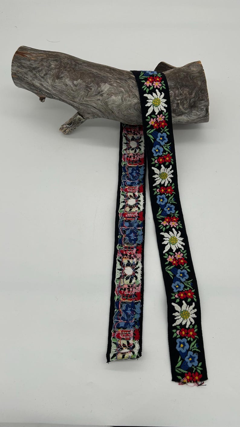 May include: Two decorative ribbons with floral patterns. One ribbon features a repeating pattern of colourful flowers on a black background. The other ribbon has a patchwork design with various colours and patterns. The ribbons are draped over a piece of driftwood.