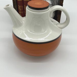 Könnte beinhalten: Eine weiß-orangefarbene Keramik-Teekanne mit schwarzer Akzentlinie. Die Teekanne hat einen runden Körper, einen gebogenen Griff und einen Ausguss. Der Deckel ist braun. Der Hintergrund umfasst eine Holzkiste, Bücher und ein Glas.