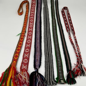 May include: Seven long, narrow woven bands or belts arranged vertically. Each features distinct geometric patterns and color combinations, including red, yellow, green, black, white, and grey. Most bands terminate in loose, multi-colored yarn tassels.