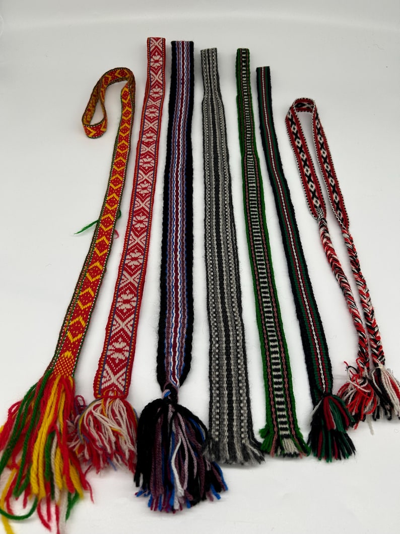 May include: Seven long, narrow woven bands or belts arranged vertically. Each features distinct geometric patterns and color combinations, including red, yellow, green, black, white, and grey. Most bands terminate in loose, multi-colored yarn tassels.