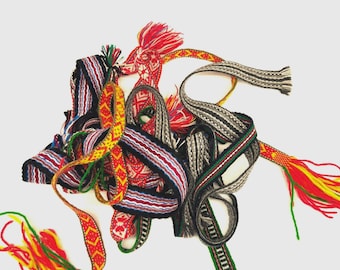 May include: A collection of woven fabric bands featuring intricate geometric patterns in vibrant colors including red, yellow, green, black, and white. These narrow, decorative textile strips are intertwined, with some ends finished with loose, multi-colored yarn tassels.