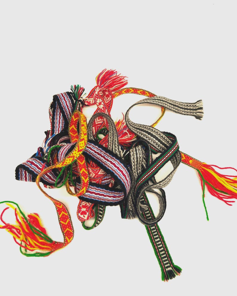 May include: A collection of woven fabric bands featuring intricate geometric patterns in vibrant colors including red, yellow, green, black, and white. These narrow, decorative textile strips are intertwined, with some ends finished with loose, multi-colored yarn tassels.