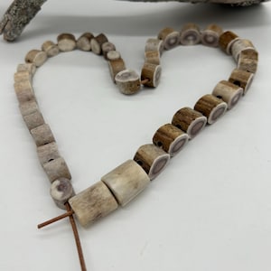 Pode incluir: Um colar feito de contas de chifre naturais e claras, enfiadas em um cordão marrom, dispostas em forma de coração. As contas variam em tamanho e forma, algumas mostrando a estrutura interna do chifre. O colar é exibido em uma superfície branca.