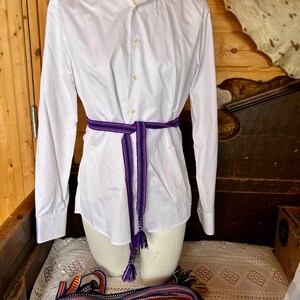 Pu&ograve; includere: Una camicia bianca a maniche lunghe &egrave; stretta in vita con una cintura intrecciata viola e nera. La cintura ha nappe decorative. Un'altra cintura intrecciata colorata &egrave; drappeggiata sotto.