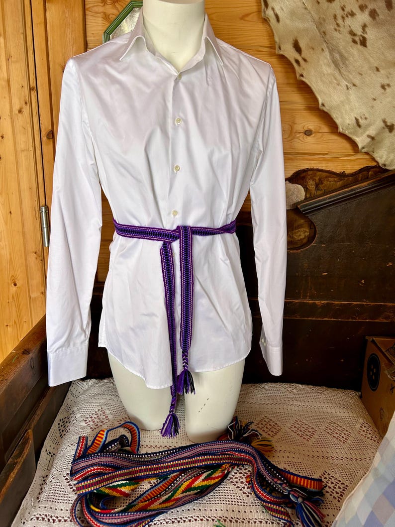 Pu&ograve; includere: Una camicia bianca a maniche lunghe &egrave; stretta in vita con una cintura intrecciata viola e nera. La cintura ha nappe decorative. Un'altra cintura intrecciata colorata &egrave; drappeggiata sotto.
