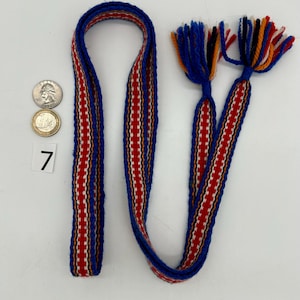 Pu&ograve; includere: Una cinghia tessuta con un motivo blu, rosso e bianco, con nappe. La cinghia presenta un motivo ripetuto di strisce verticali rosse e bianche. Le nappe sono blu, arancioni, rosse e bianche. Il numero 7 &egrave; stampato su un quadrato bianco.