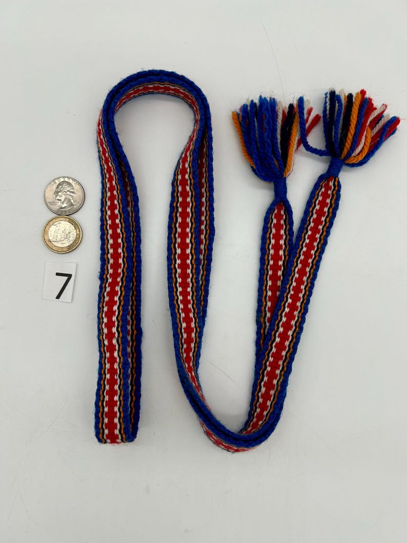 Pu&ograve; includere: Una cinghia tessuta con un motivo blu, rosso e bianco, con nappe. La cinghia presenta un motivo ripetuto di strisce verticali rosse e bianche. Le nappe sono blu, arancioni, rosse e bianche. Il numero 7 &egrave; stampato su un quadrato bianco.