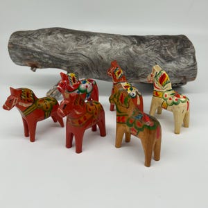 Pode incluir: Uma coleção de seis cavalos Dala pintados à mão em várias cores, incluindo vermelho, amarelo e tons de madeira natural. Os cavalos de madeira são decorados com padrões florais tradicionais e estão sobre uma superfície branca, com um pedaço de madeira flutuante no fundo.