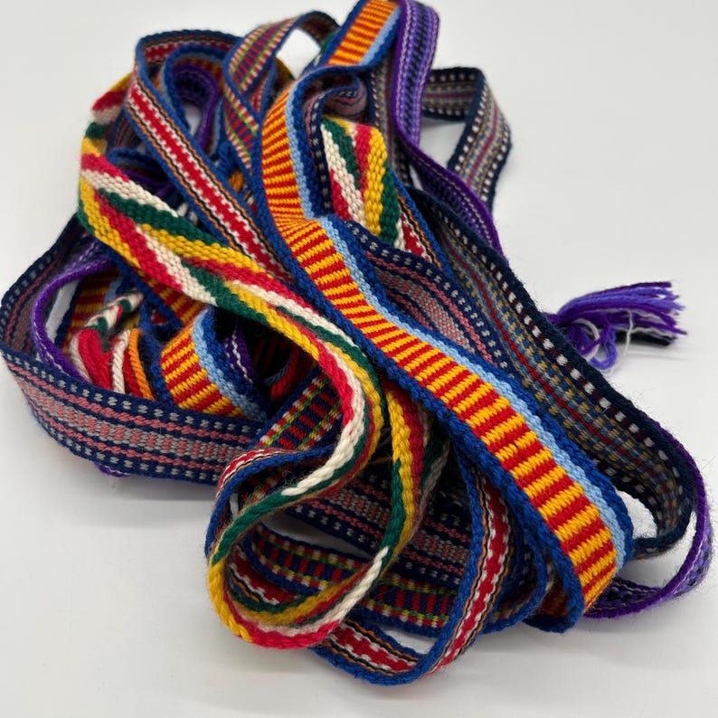 Pu&ograve; includere: Una collezione di nastri tessuti colorati con vari motivi. I nastri presentano strisce e disegni geometrici in rosso, giallo, blu, verde e bianco. Alcuni nastri hanno una frangia di fili viola.
