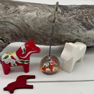 May include: A collection of Dala horse-themed items. Includes a red and white painted wooden horse, a clear sphere with a miniature horse inside, a white horse figurine, and a red horse-shaped clip.