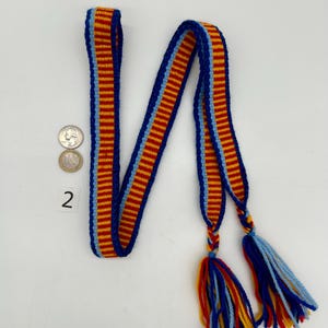 Pu&ograve; includere: Una cinghia tessuta con un motivo ripetuto di strisce rosse, gialle e blu. La cinghia ha bordi blu e termina con nappe rosse, gialle e blu. Sono visibili due monete e il numero 2.