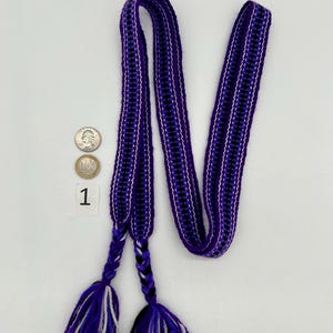Pu&ograve; includere: Una cintura intrecciata nei toni del viola, nero e bianco, con nappe intrecciate alle estremit&agrave;. La cintura &egrave; esposta su sfondo bianco. La cintura &egrave; lunga circa 152 cm.