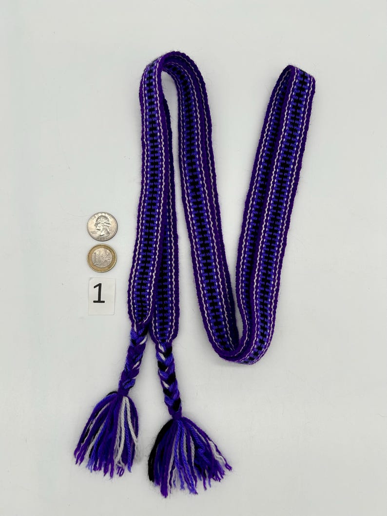 Pu&ograve; includere: Una cintura intrecciata nei toni del viola, nero e bianco, con nappe intrecciate alle estremit&agrave;. La cintura &egrave; esposta su sfondo bianco. La cintura &egrave; lunga circa 152 cm.