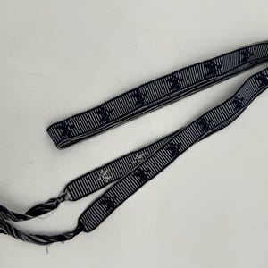 May include: A navy blue and white striped ribbon with a repeating pattern of a stylized emblem. The ribbon is folded and has frayed ends. The ribbon is lying on a white surface.